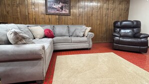 Un salón con un sofá seccional gris, un sillón de cuero negro, una alfombra roja estampada y una pared de paneles de madera con un cuadro enmarcado.