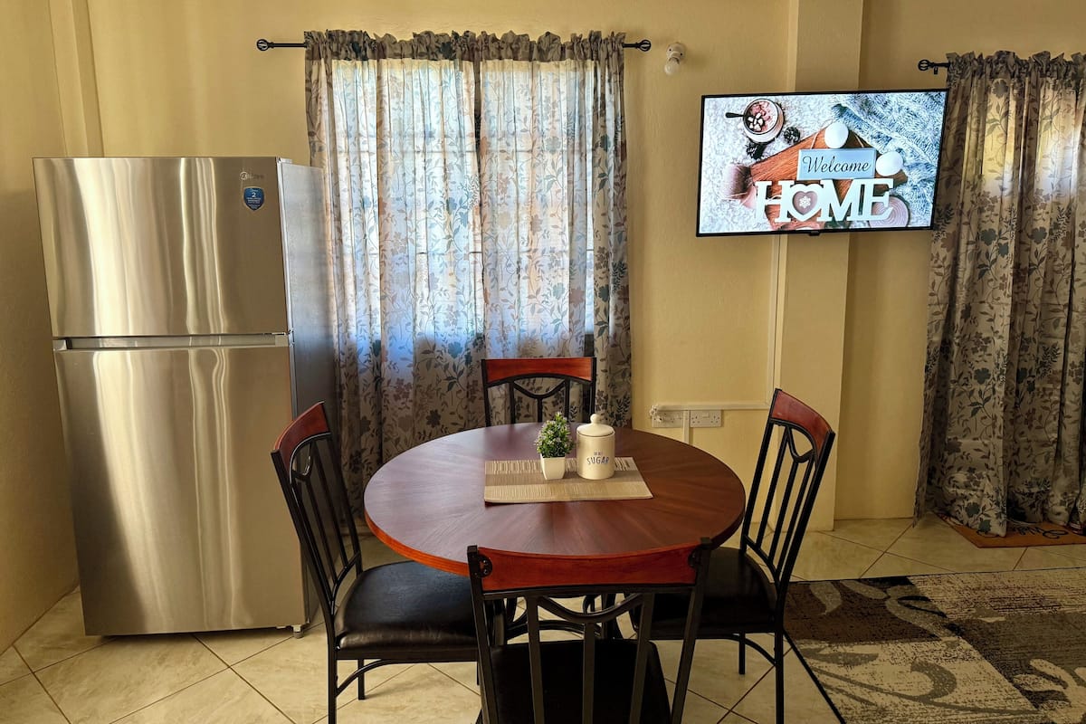 A compact kitchen with a stainless steel refrigerator, a round wooden dining table with four chairs, and a flat-screen TV mounted on the wall displaying a welcome message.