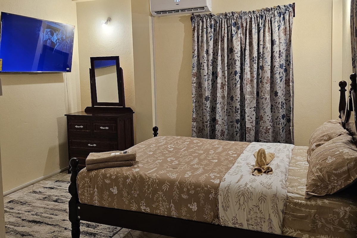 A bedroom with a bed, a flat-screen TV, an air conditioner, and a dark wooden dresser.