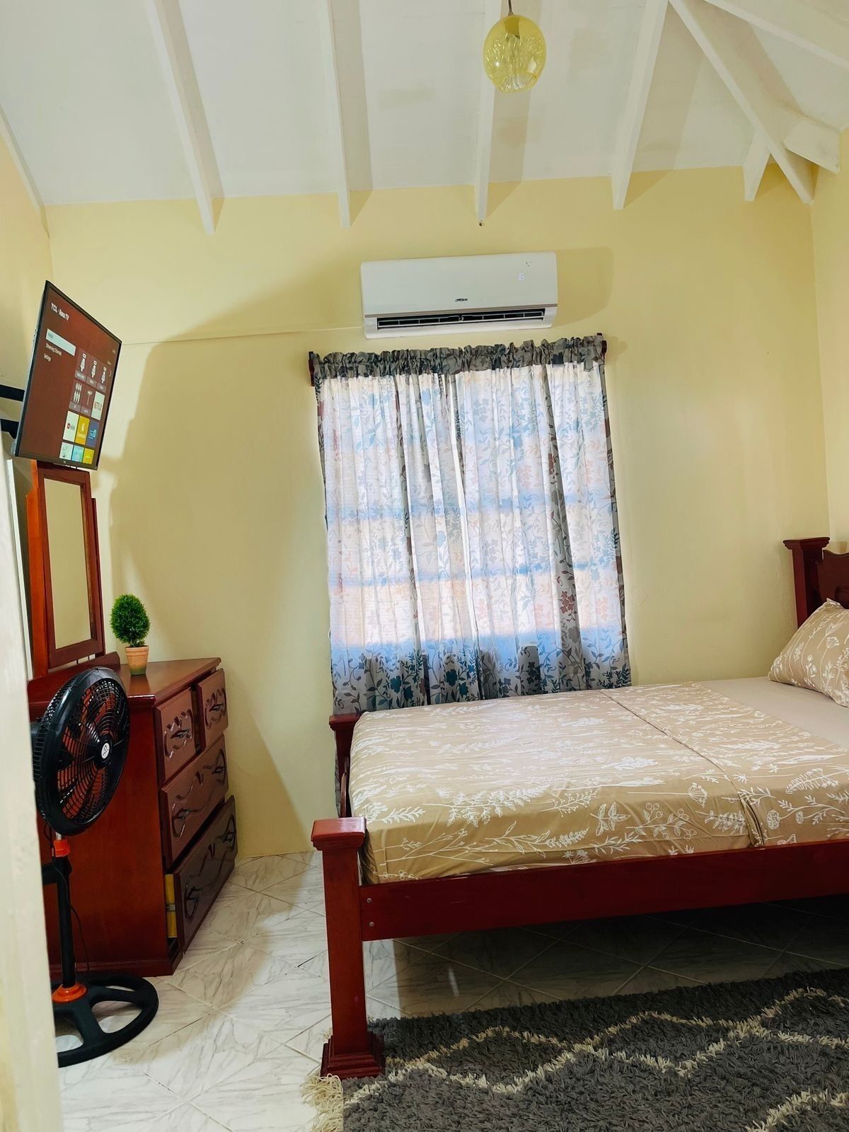 A bedroom with a bed, a dresser, a fan, an air conditioner, and a window with floral curtains.