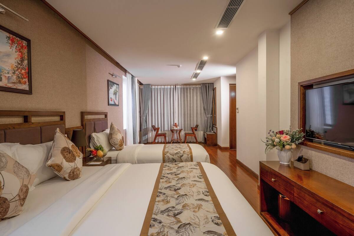 Ein Hotelzimmer mit zwei Betten, einem Fernseher, einem Schreibtisch mit Blumenvase und einem Essbereich mit Stühlen und Vorhängen.