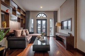 A modern living room with a sofa, a coffee table, and a TV mounted on the wall. There is a bookshelf with decorative items and a large window with curtains. - Canvas Sapa Legend Hotel (Ho Chi Minh Municipality)