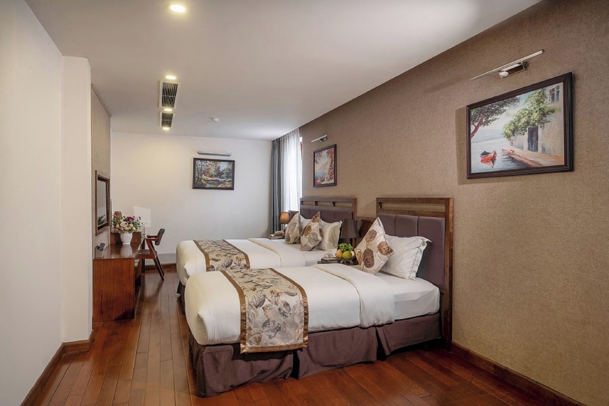 Een hotelkamer met twee bedden, een bureau en een stoel. Er hangen foto’s in lijsten aan de muren en een raam met gordijnen.