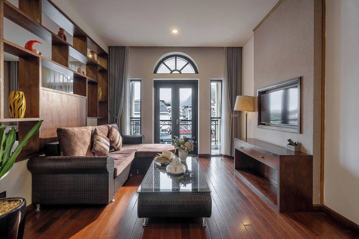 Ein modernes Wohnzimmer mit einer Couch, einem Couchtisch und einem an der Wand befestigten Fernseher. Ein Bücherregal mit Dekorationselementen und ein großes Fenster mit Vorhängen.