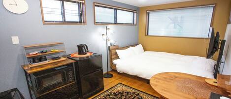 Habitación de hotel compacta con cama, kitchenette pequeña, cafetera, escritorio con computadora y dos ventanas con persianas.