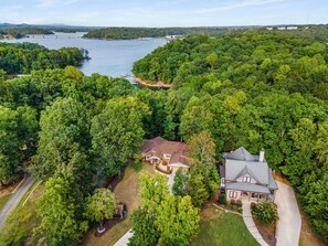 An aerial view of a residential area with a large house surrounded by dense forest, a dock, and a lake in the background. - Cozy 4bd lakefront home in Lake Lanier w/ HotTub, FirePit, Private dock, Wifi (Gainesville)