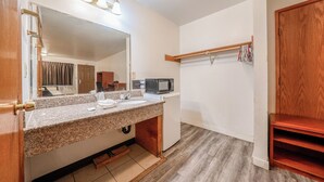 A bathroom with a granite countertop sink, a mirror, a vanity, a microwave, and a wooden shelf.