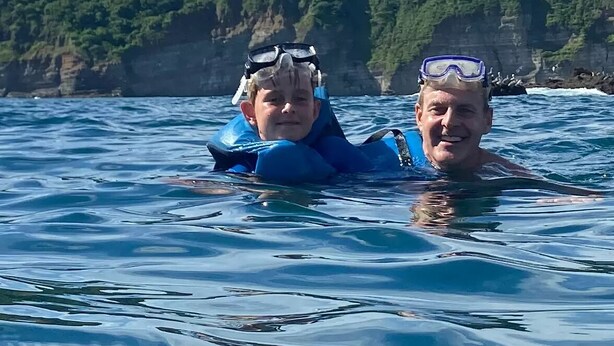Two people in blue wetsuits and goggles floating in calm water with a rocky coastline and greenery in the background. - Hotel Ingigante Poolside Room (El Gigante)