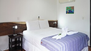 Hotellihuone, jossa on sänky, yöpöytälamppu, yöpöytä, ilmastointilaite ja seinälle ripustettu taulu.