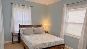 Ein Schlafzimmer mit einem hölzernen Bett, einem Nachttisch, einer Lampe und einem Fenster mit Jalousien.