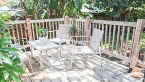 A wooden deck with white outdoor furniture, including a table and two chairs, surrounded by a wooden fence and lush greenery.