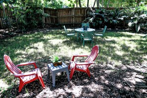 Un patio trasero con una mesa de madera y dos sillas rojas, rodeado de abundante vegetación y una valla de madera.