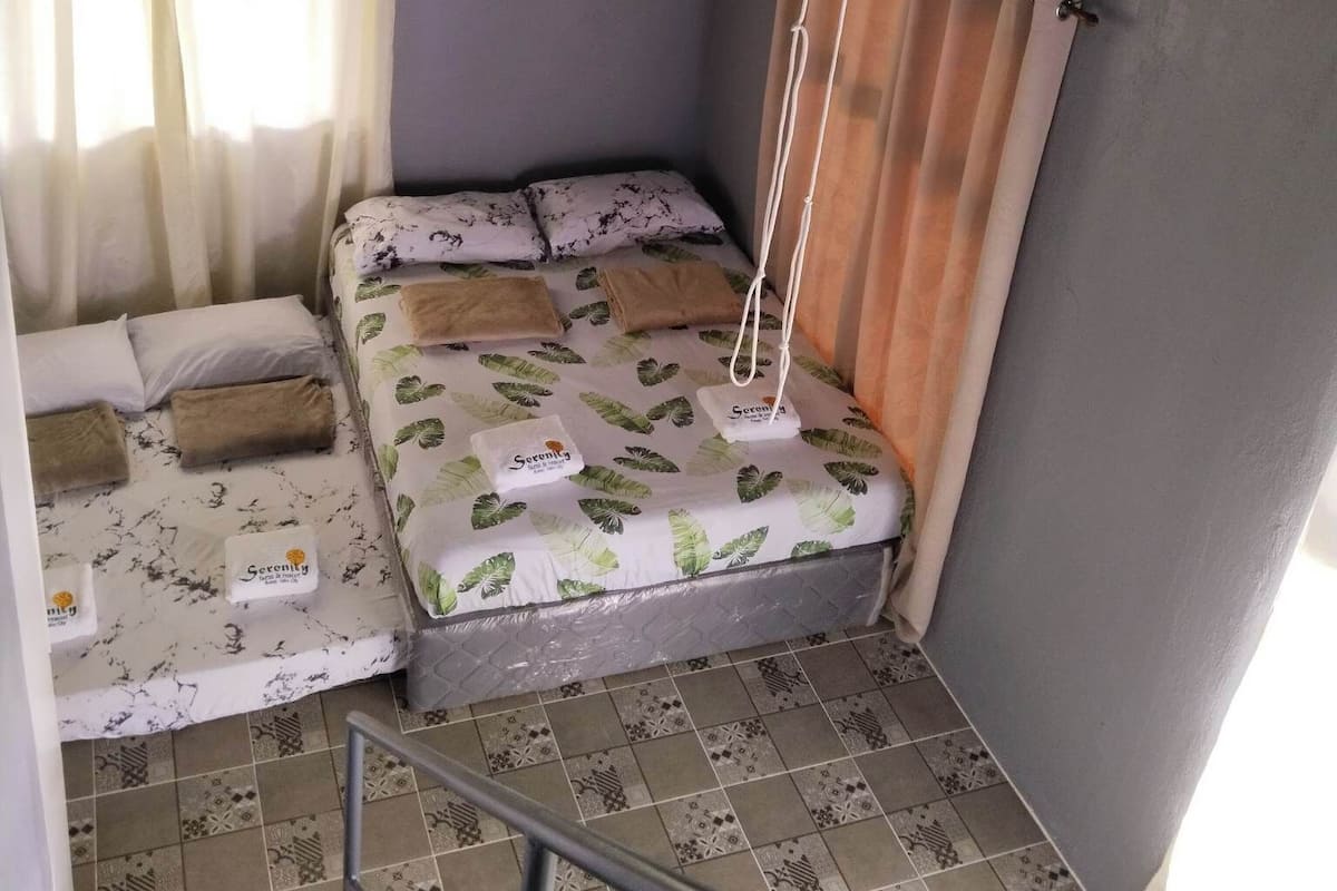 Une petite pièce, de taille modeste, comprenant un lit, un ventilateur de plafond et une fenêtre avec des rideaux.
