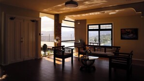 Een ruime kamer met een hoge plafond, een plafondventilator, een groot raam met uitzicht op bergen, en een zithoek met een tafel en stoelen.