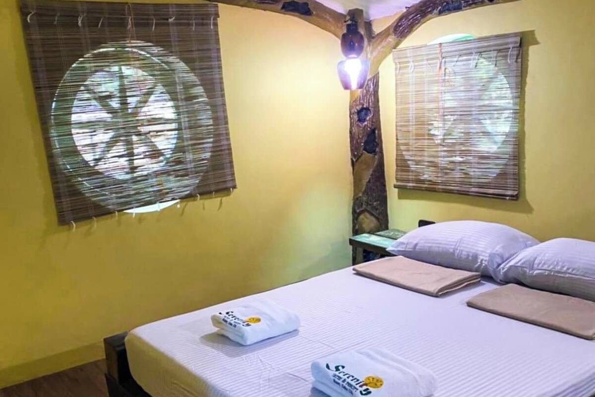 Un lit bien fait, avec des oreillers et une couverture, une petite table avec une lampe, et deux fenêtres équipées de stores.