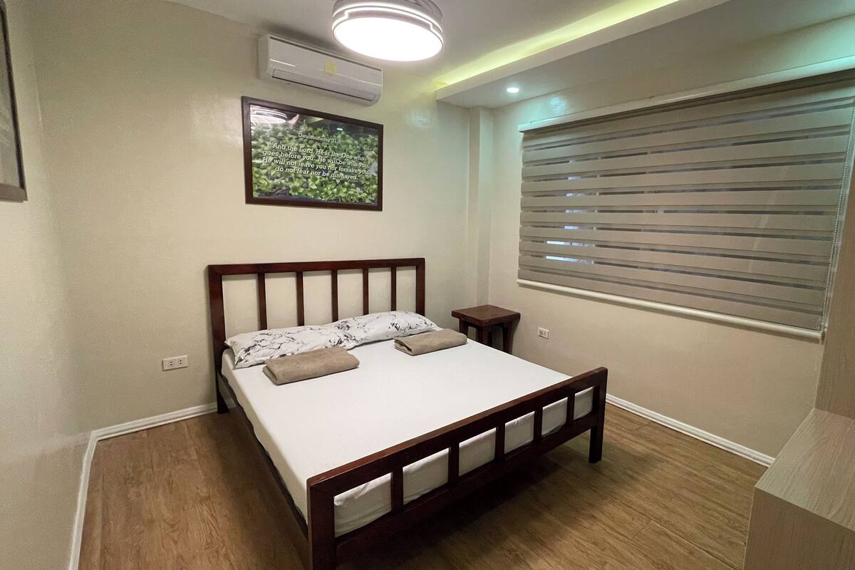 Une chambre à coucher avec un lit en bois, une petite table de chevet, un climatiseur fixé au mur et une fenêtre avec des stores.