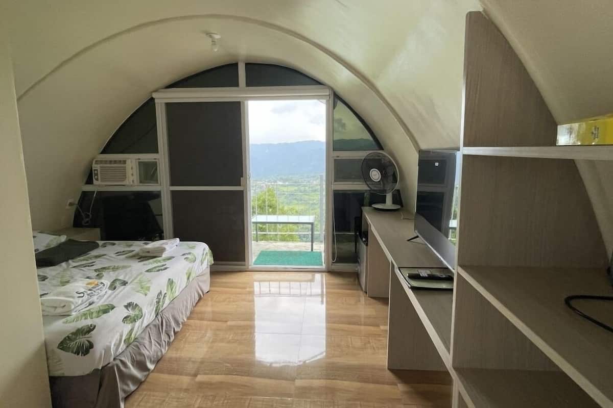 Une petite pièce comprenant un lit, un bureau, une chaise, un ventilateur et une fenêtre donnant sur les montagnes.