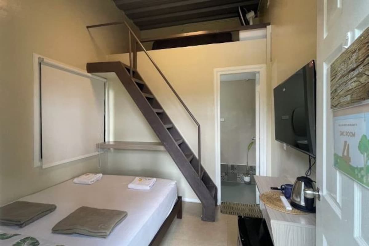 A compact hotel room with a bed, a small kitchenette, a television, and a staircase leading to an upper level.