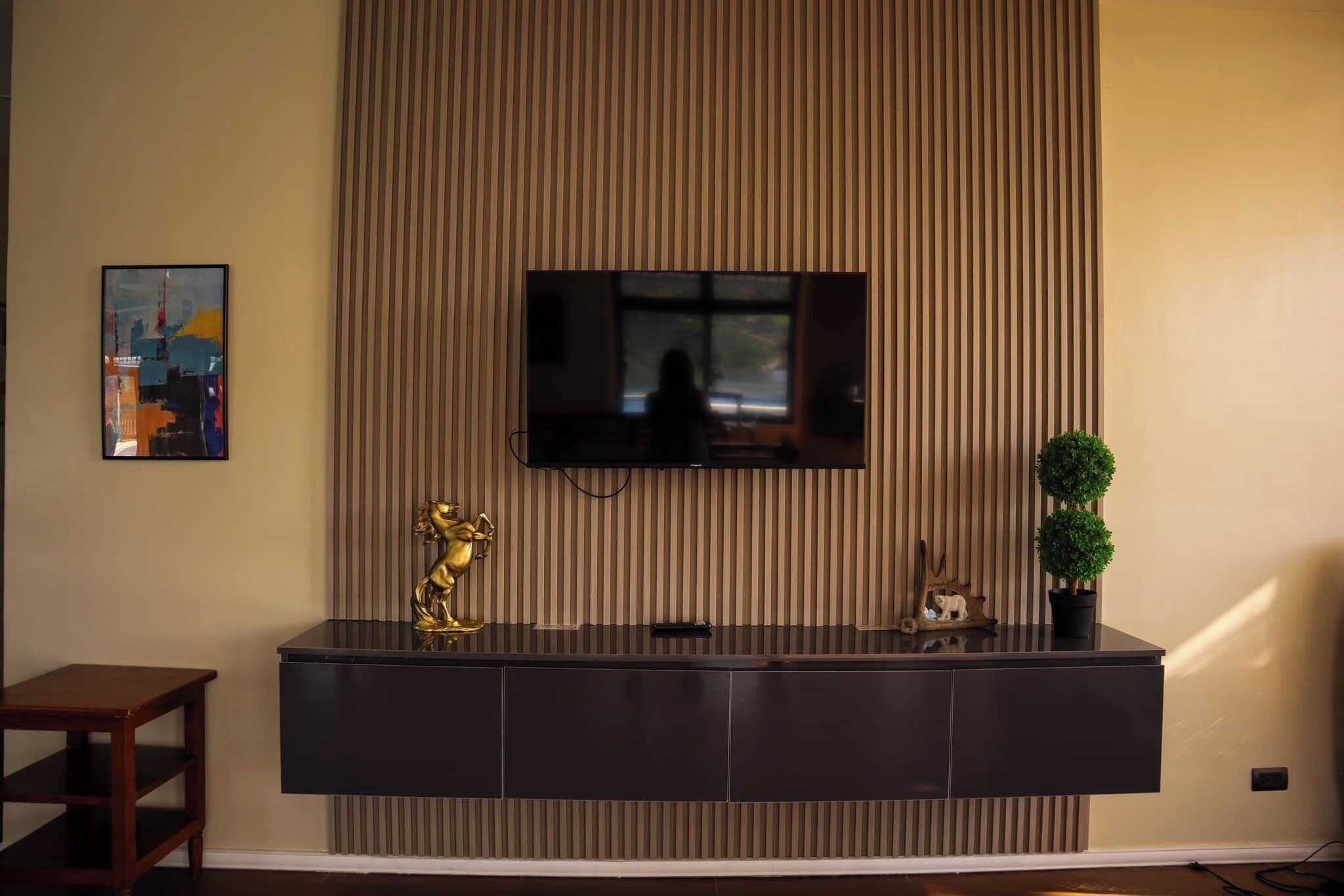 A modern living room with a large flat-screen TV mounted on a wall with vertical blinds, a dark wooden console table with decorative items, and a small potted plant.