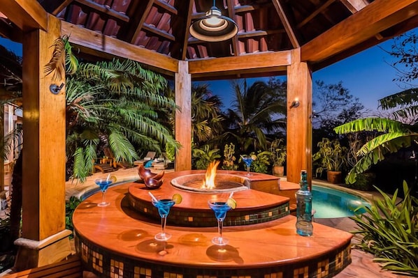 A wooden outdoor bar area with a fire feature, surrounded by tropical plants and a pool.