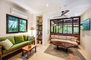 A modern living room with a green sofa, a wooden coffee table, and a beige sofa. There are large windows with a view of greenery outside.