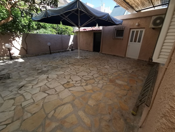 A stone patio with an umbrella, a door, and a wall with a window. - RENT HOUSE ROOMS KALAMATA (Agios Dionisios)
