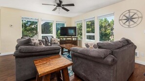 A spacious living room with a ceiling fan, large windows, a flat-screen TV, and a rustic wooden coffee table. - Makena's -near beach, game room, hot tub, dog ok (Pacific City)