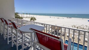 A balcony with white and red chairs, a small table, and a view of a beach and ocean. - Beachfront Oasis with Dual Balconies, Pool, Hot Tub & Stunning Sunset Views in Madeira Beach (Madeira Beach)
