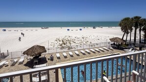 A beach view from a balcony with a pool, lounge chairs, and a thatched umbrella. - Beachfront Oasis with Dual Balconies, Pool, Hot Tub & Stunning Sunset Views in Madeira Beach (Madeira Beach)