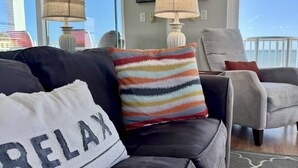 A living room with a black leather sofa, a colorful striped pillow, and a white pillow with "RELAX" on it. There are two lamps on the table, a grey armchair, and a view of the ocean through large windows. - Beachfront Oasis with Dual Balconies, Pool, Hot Tub & Stunning Sunset Views in Madeira Beach (Madeira Beach)