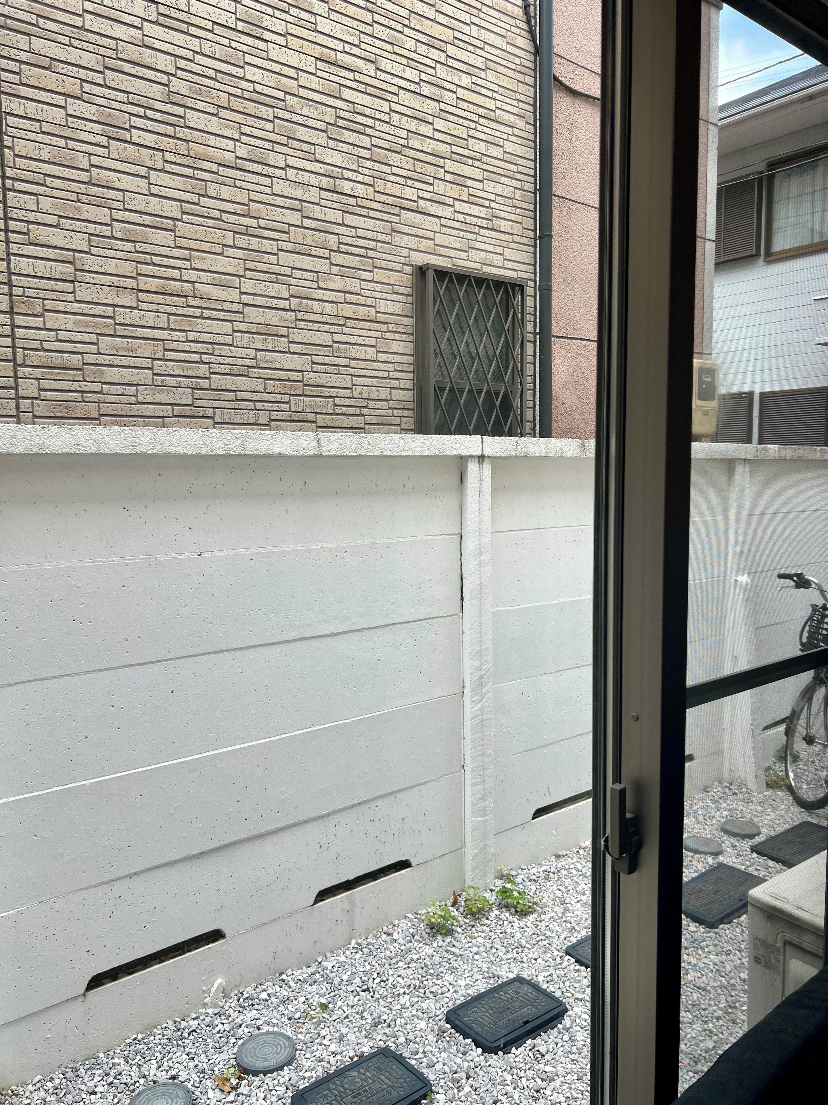 A view through a glass door of a building with a brick exterior, a concrete wall, and a gravel ground with a bicycle in the background.