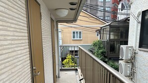 A narrow corridor with a door on the left, a balcony with a metal railing on the right, and a building with windows and air conditioning units in the background. - Gracious Higashi Shinjuku (Tokyo)