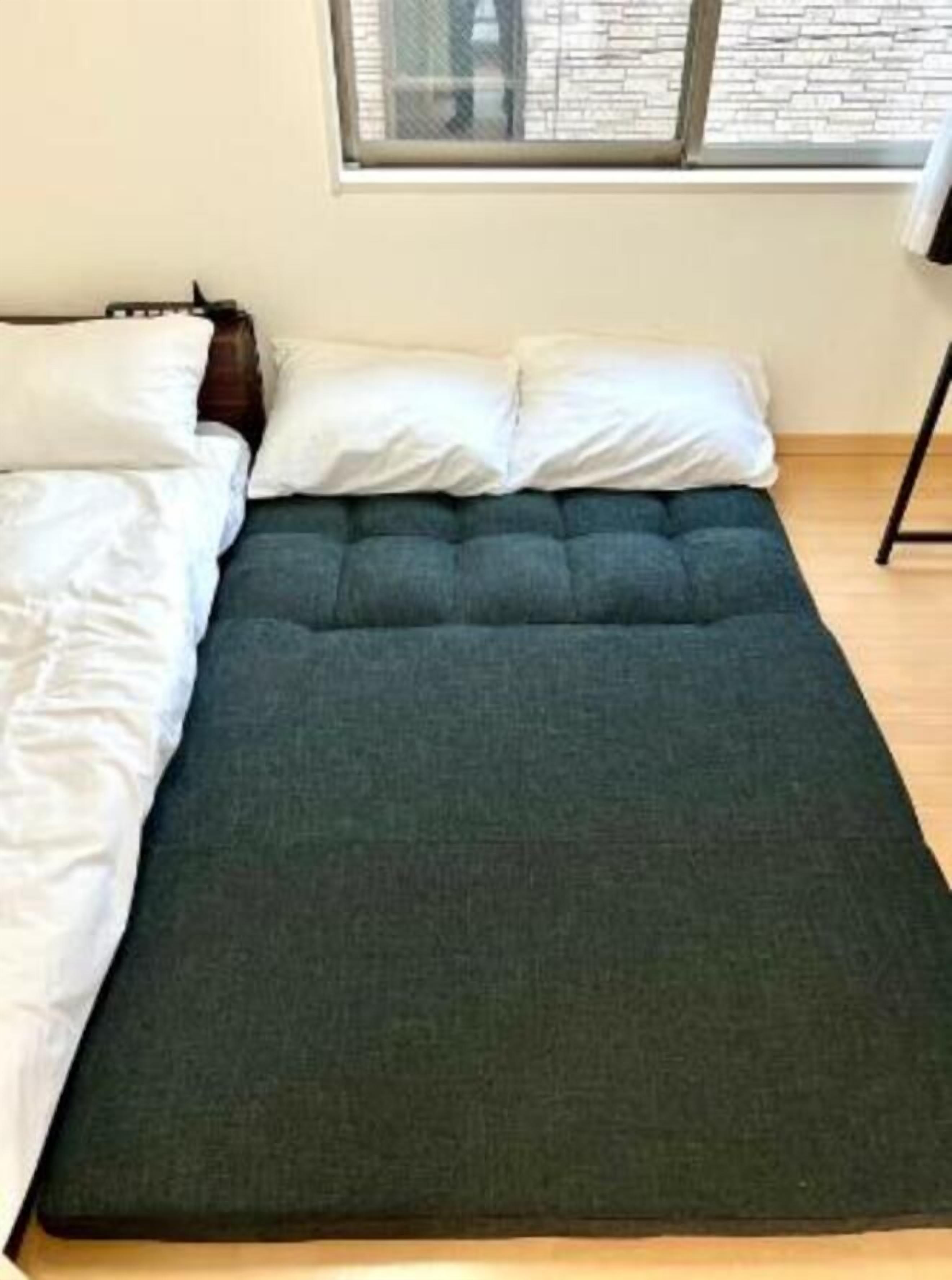 A bed with a dark mattress and white pillows, a window with a view of a brick wall, and a wooden floor.