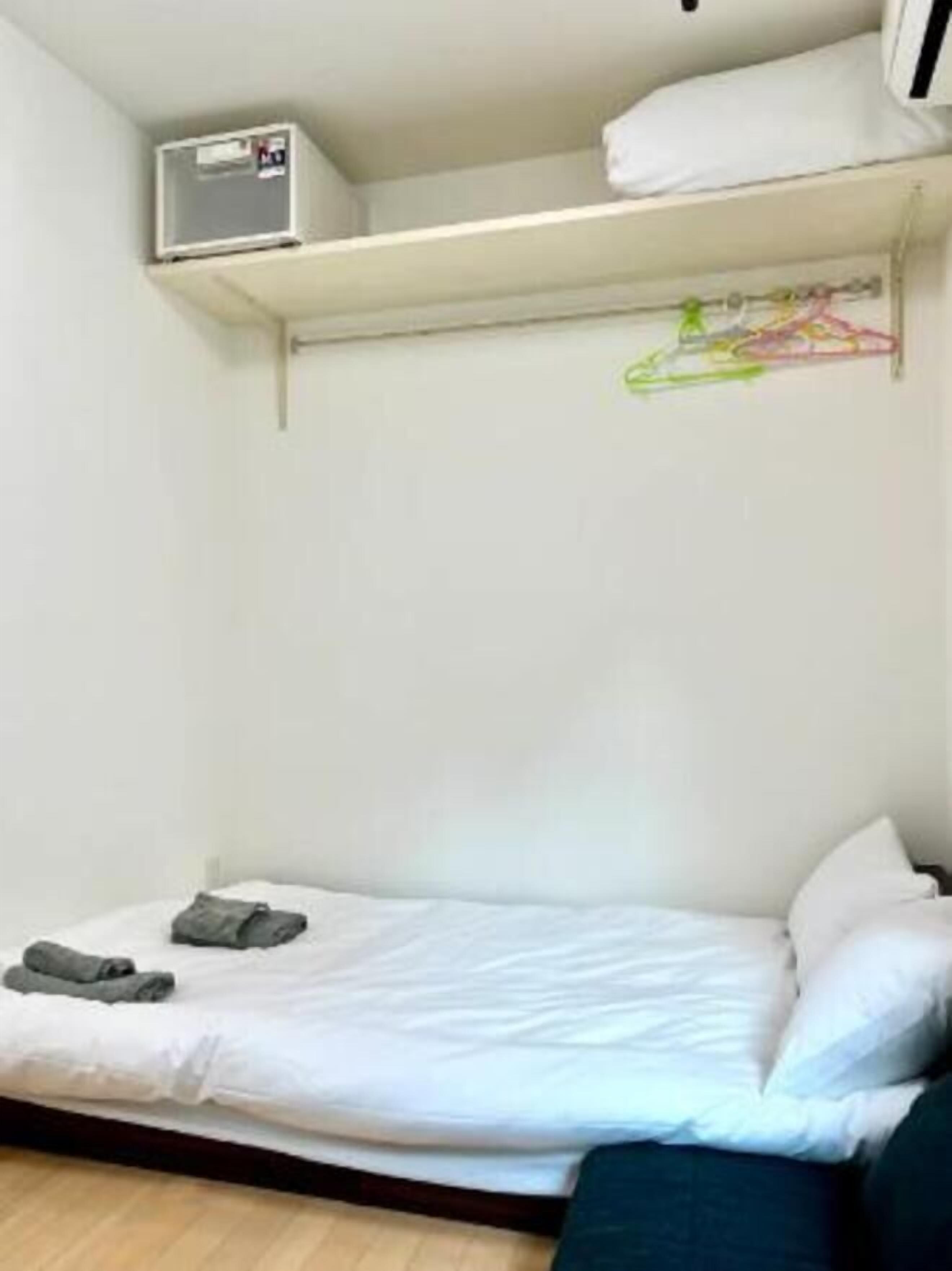 A small, single-bed room with a white mattress, a pair of folded clothes on the bed, a small shelf with a white object, and a wall-mounted air conditioning unit.