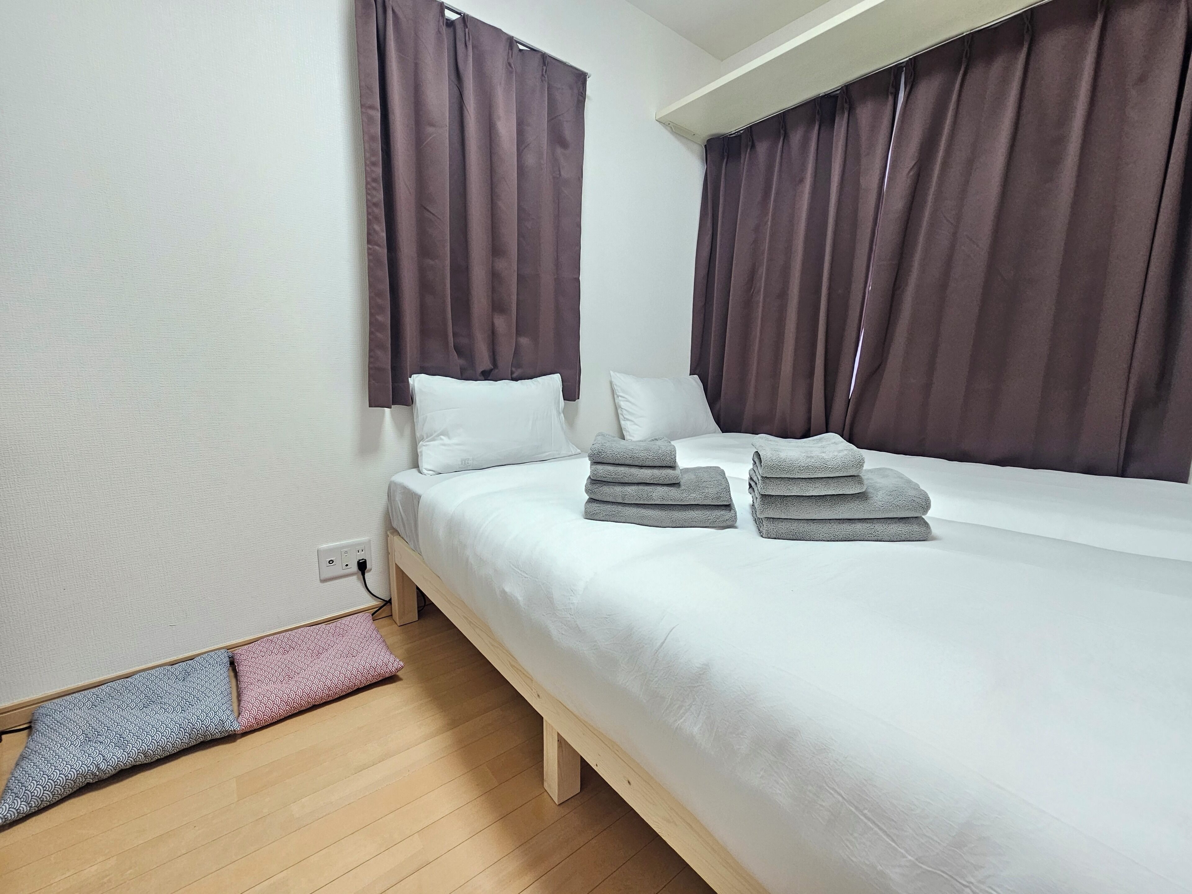 A neatly made bed with pillows and folded towels, a wooden floor, and brown curtains.