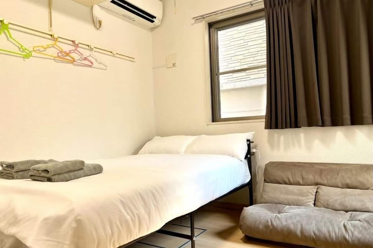 A compact hotel room with a bed, a sofa, a window with curtains, an air conditioning unit, and a wall-mounted light fixture.