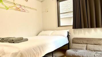 A compact hotel room with a bed, a sofa, a window with curtains, an air conditioning unit, and a wall-mounted light fixture.