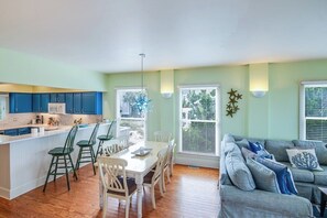 A modern kitchen with blue cabinets, a dining area with a white table and green chairs, and a living room with a gray sofa and blue cushions.