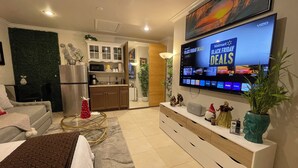 A modern living room with a flat-screen TV displaying Black Friday deals, a kitchen area with white cabinets, a dining table, and a sofa. - Elegant Tranquil Restful Oasis with Pure Serenity Pool and Garden Suite Studio (Miami)