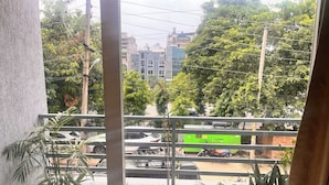 Un balcón con plantas en macetas, vistas a una calle con coches y un autobús verde, y edificios al fondo.