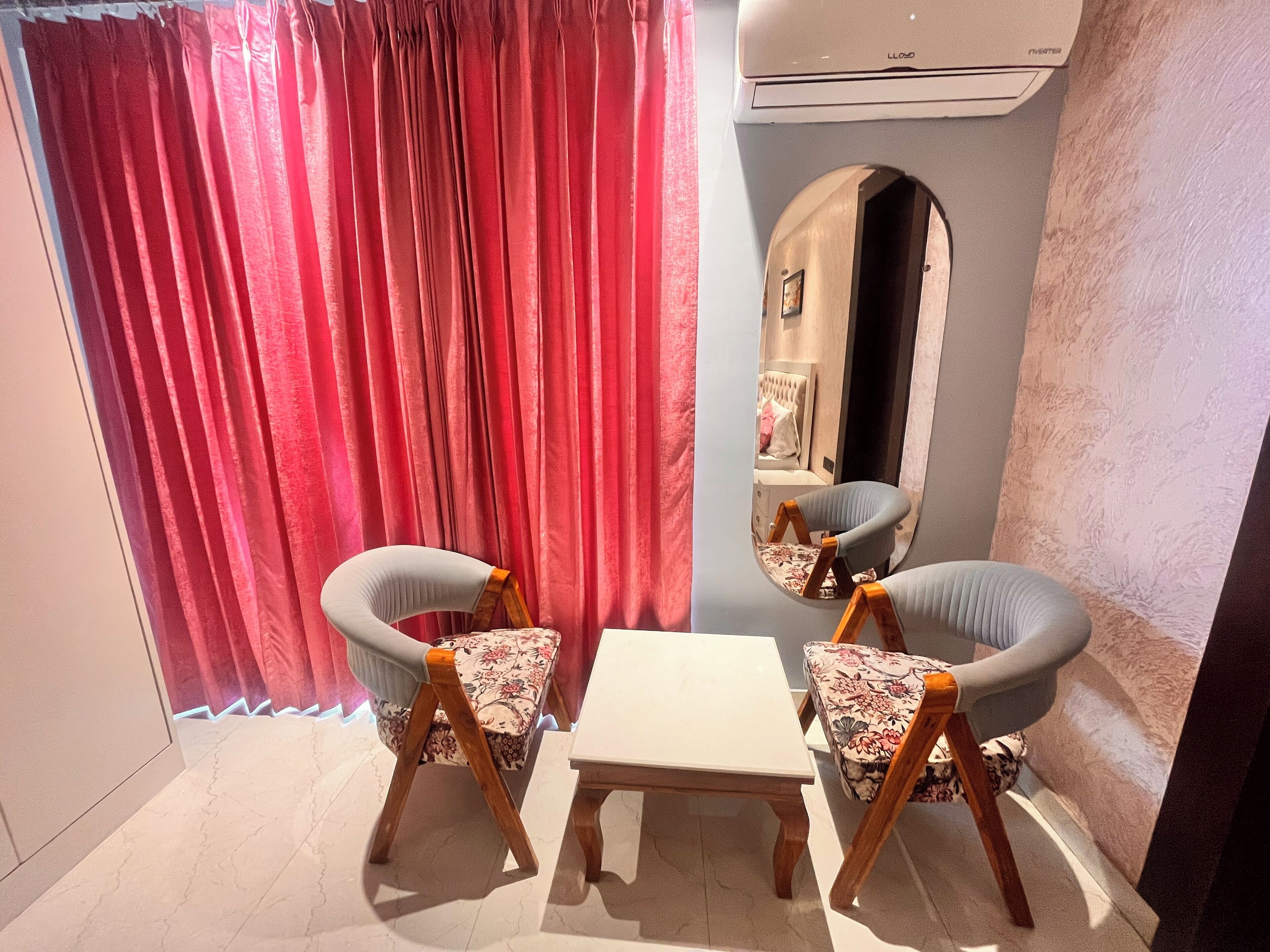 A room with a white table and two wooden chairs with floral upholstery. A red curtain and a mirror with a picture frame are visible.
