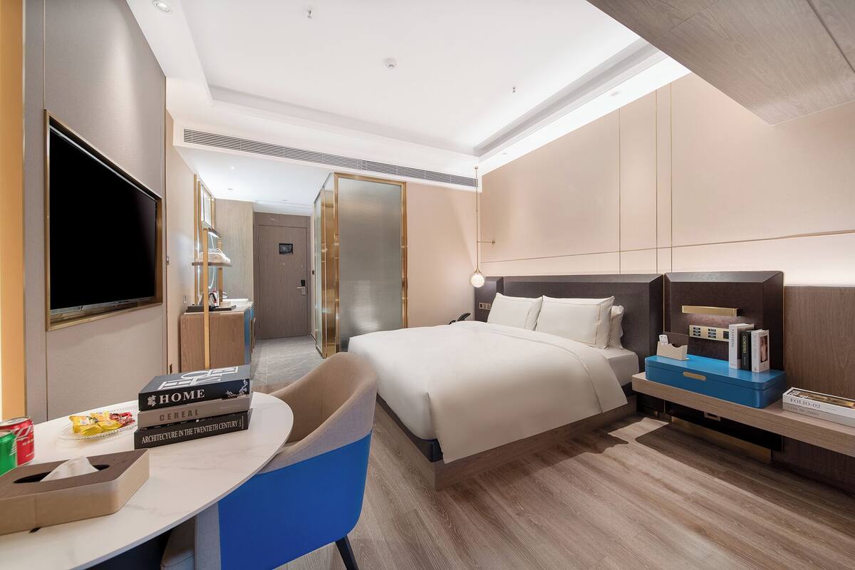 Quarto de hotel moderno com uma cama grande, uma escrivaninha com cadeira, uma TV e uma mesa de cabeceira com livros.