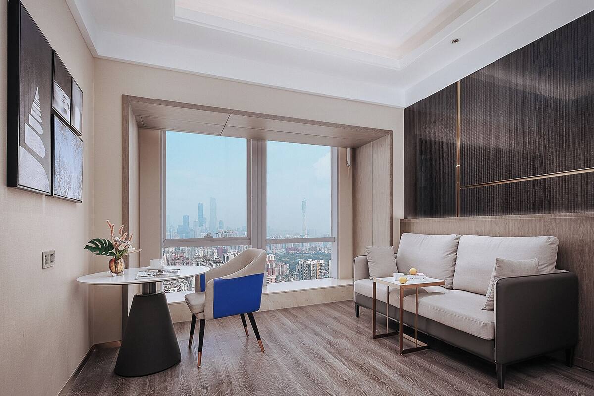 Uma sala de estar moderna com sofá, mesa de centro e escrivaninha com cadeira. Há uma janela grande com vista para a cidade.