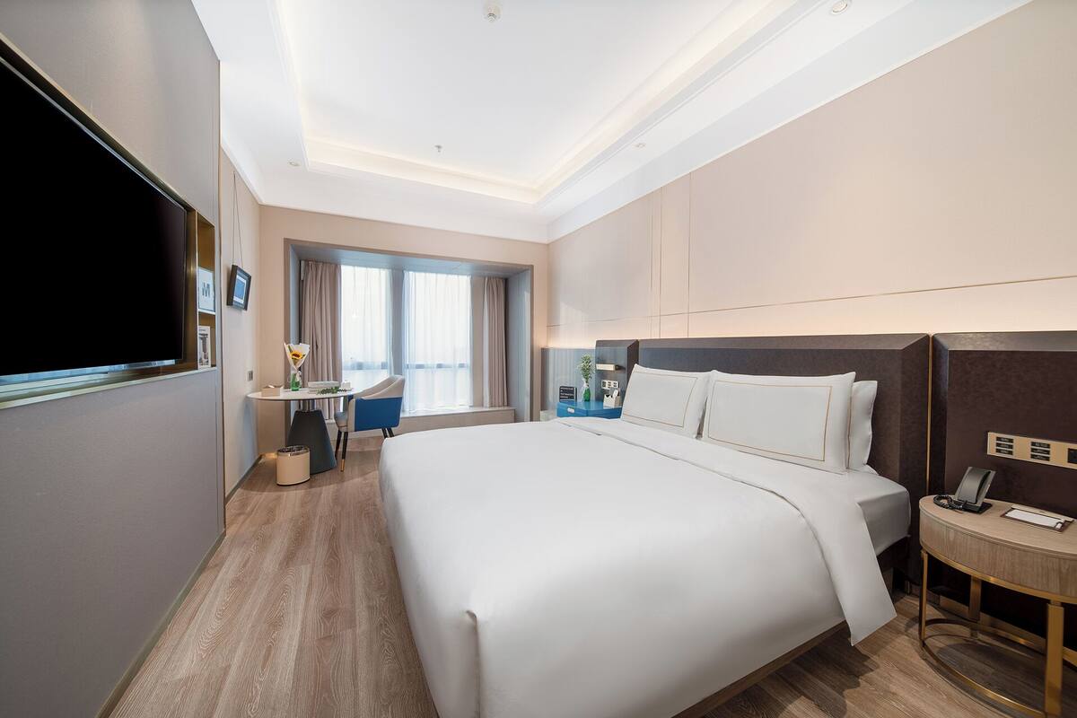 Quarto de hotel com uma cama grande, uma escrivaninha com cadeira, televisão e mesa de cabeceira com telefone.
