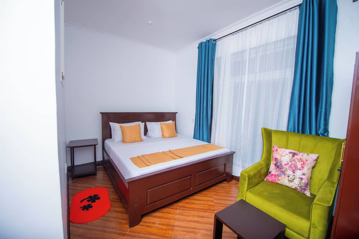 A hotel room with a bed, a green armchair, a small table, and a red mat.
