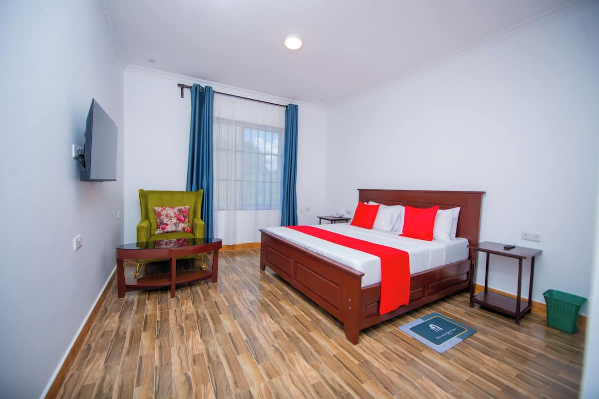 A hotel room with a wooden floor, a bed with red and white bedding, a green armchair, a small table, and a TV mounted on the wall.