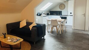 Una sala moderna con un sofá oscuro, una mesita de centro pequeña y una zona de comedor con una mesa y sillas blancas. Se ve una cocina al fondo con armarios blancos y un reloj grande en la pared.
