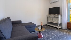 A living room with a grey sectional sofa, a small round table with a bottle and glasses, a flat-screen TV on a white cabinet, and a window with curtains. - Les Voiles by Interhome (Six Fours)