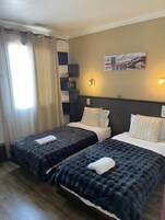 Une chambre d’hôtel avec deux lits, une table de chevet, une lampe, une étagère avec quelques objets et une affiche au mur.