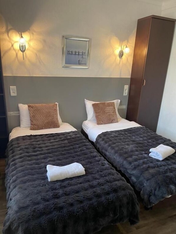 Une chambre d’hôtel avec deux lits, chacun recouvert d’une couverture épaisse et sombre et agrémenté d’un oreiller blanc. Des lampes murales sont fixées au-dessus des lits, et un tableau encadré orne le mur.
